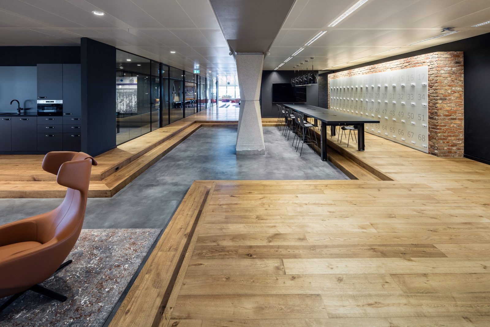 De gedeelde ruimte met schuine betonnen kolommen, houten platforms, een donkere keuken en een hoge tafel voor een genummerde wand.