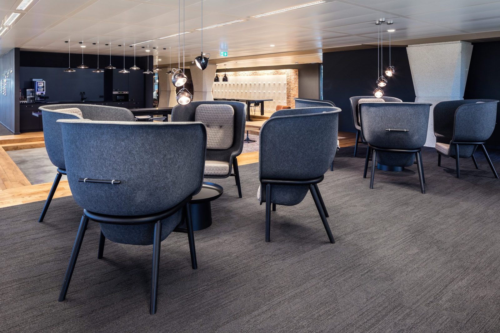 Blauwgrijze gestoffeerde stoelen rond kleine tafels, met een donkere bar en hanglampen op de achtergrond.