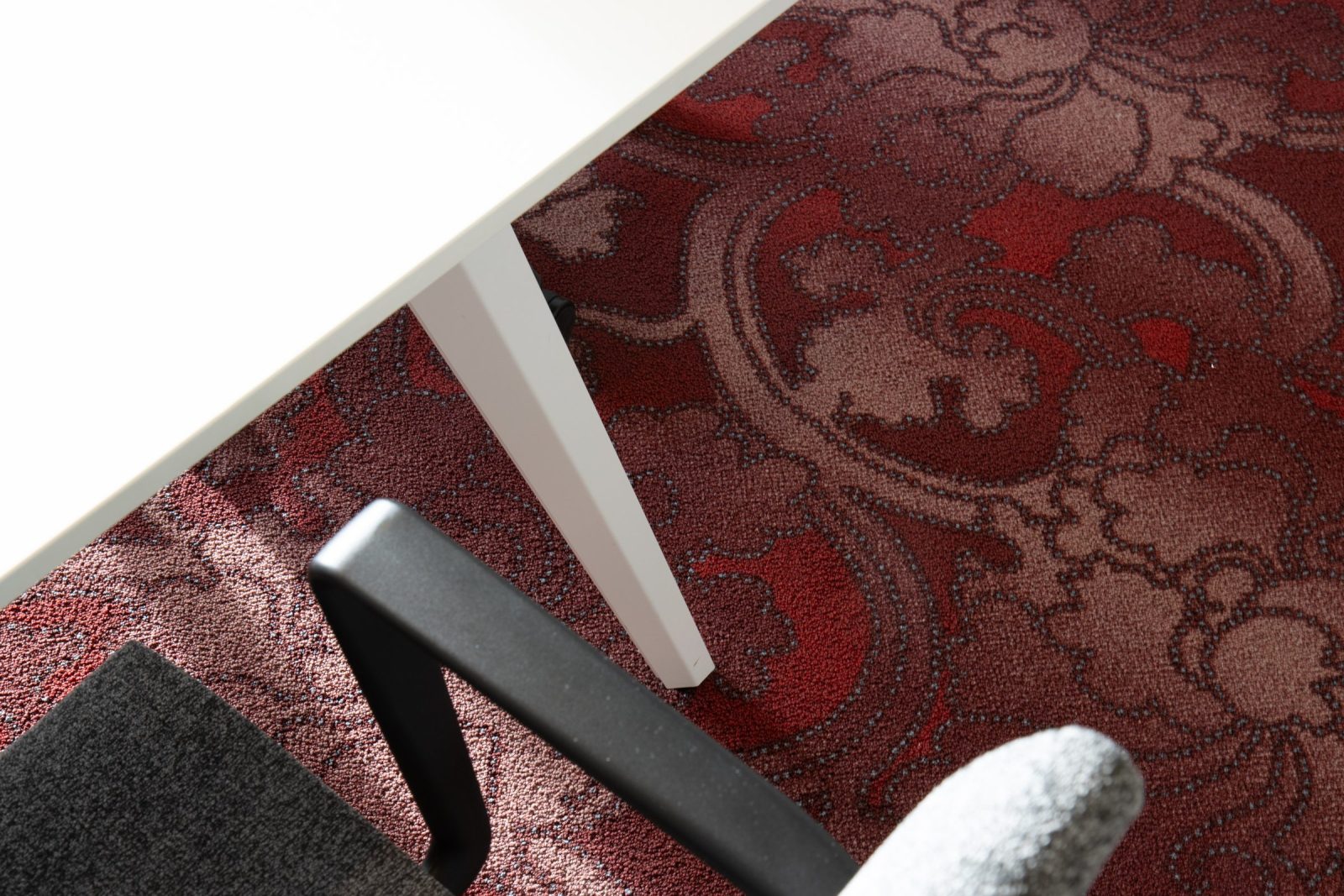 Detail van een rood tapijt met een sierlijk patroon onder een witte tafel en grijze stoel bij AVAS.