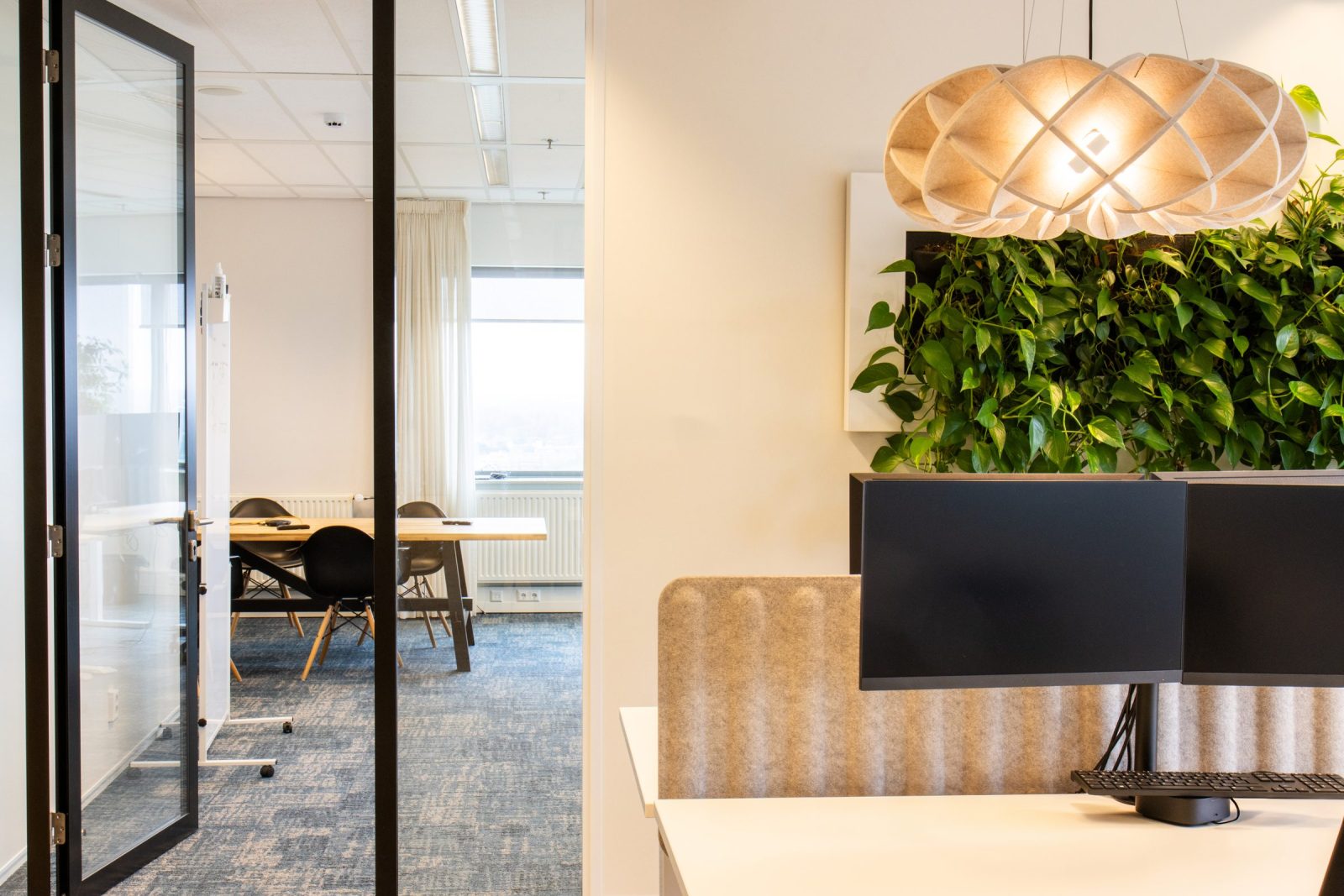 Een bureau naast planten en een houten hanglamp, met glas en licht gordijn op de achtergrond bij CLCC.