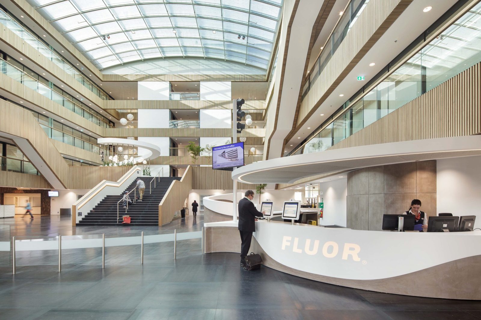 Het hoge atrium met glazen dak, verdiepingsgalerijen, een centrale trap en een witte ontvangstbalie met Fluor-logo.