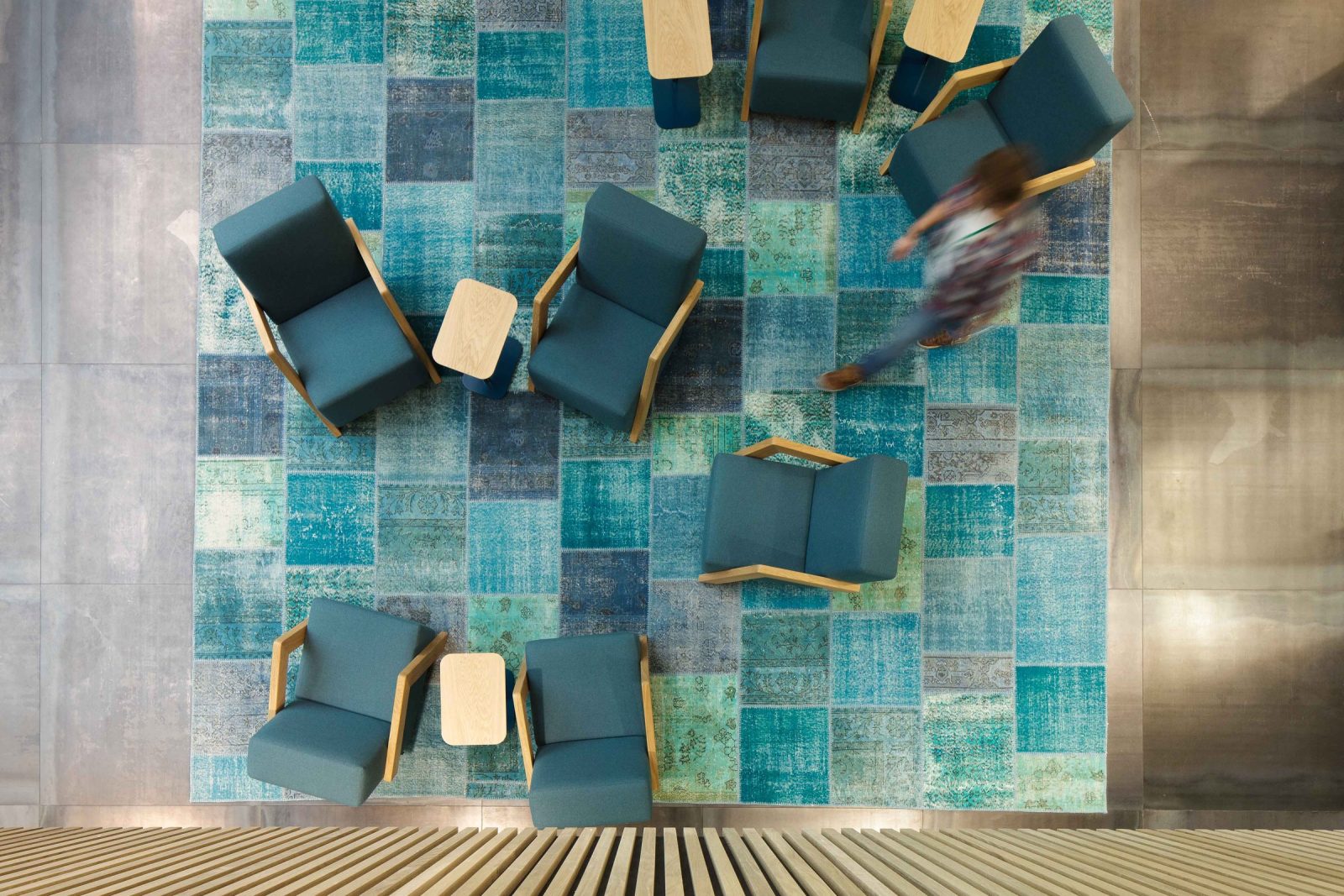 Bovenaanzicht van blauwe fauteuils en houten bijzettafels op een blauwgroen tapijt met een rechthoekig patroon.