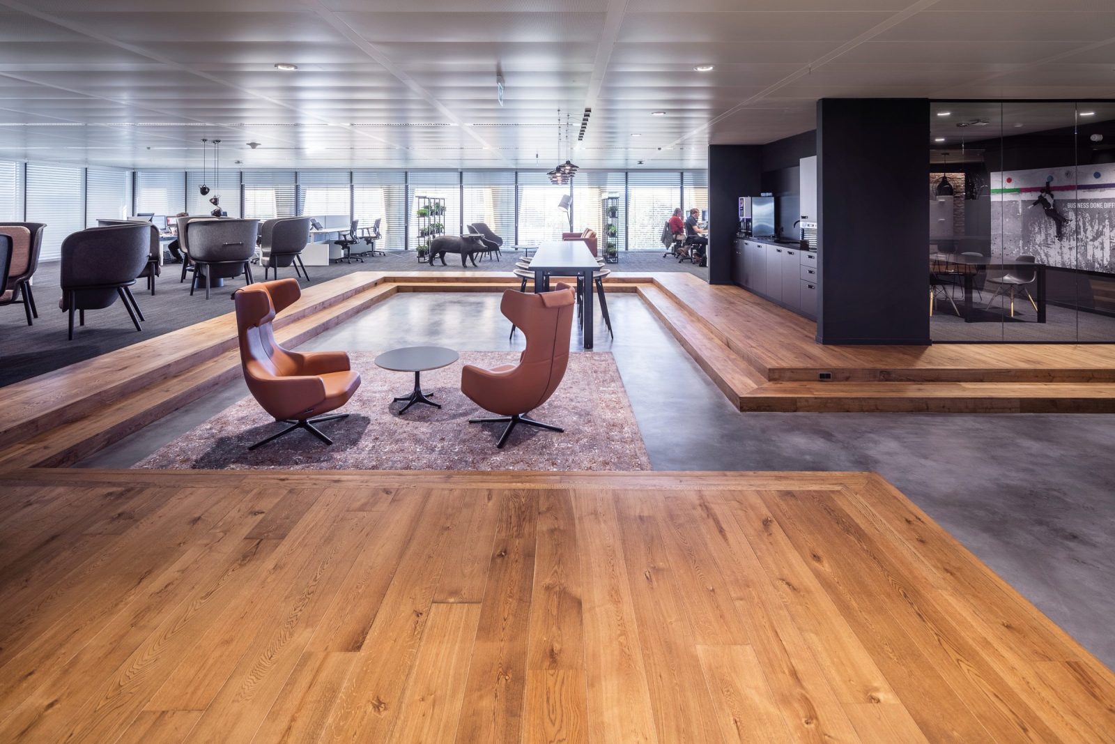 Een brede gedeelde ruimte met houten vloeren, cognackleurige stoelen, een tafel op tapijt en donkere keukenmeubels.