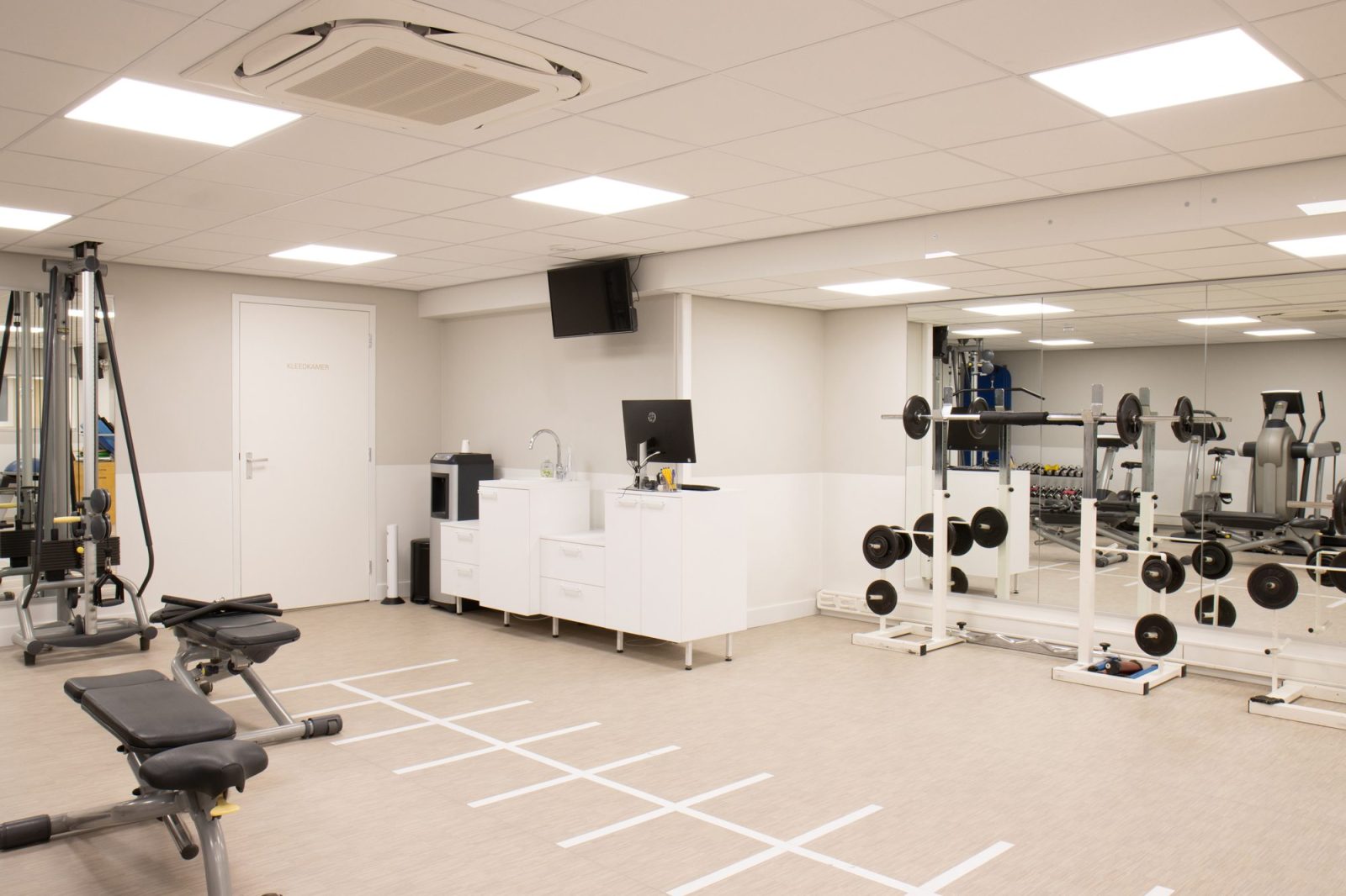 Een oefenruimte met trainingsapparatuur, gewichten, een lichte vloer met lijnen en een witte balie.