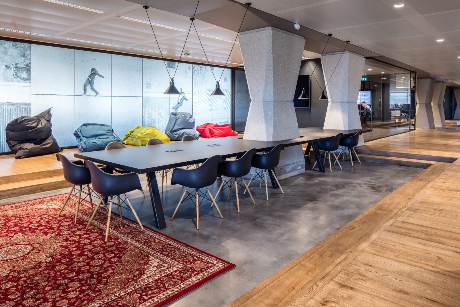 Een lange donkere tafel met stoelen naast schuine kolommen, rode vloerkleden en een verhoogde zitplek met gekleurde kussens.