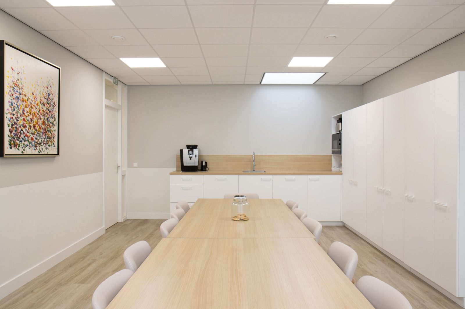Een lange houten tafel met witte stoelen in een lichte ruimte met keukenmeubels en een klein schilderij.