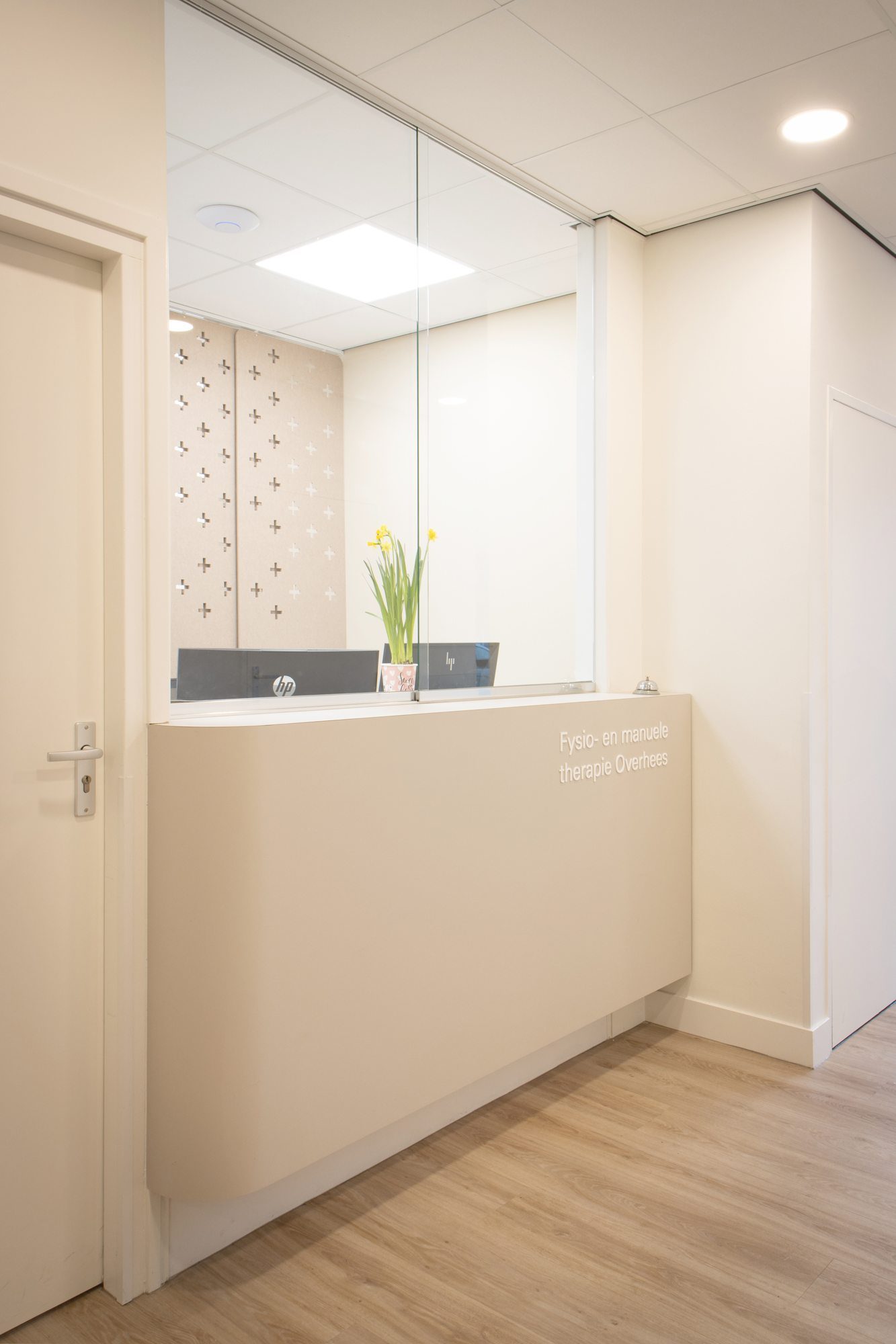Een ontvangstbalie achter een glazen wand met matte horizontale stroken, naast houten vloer en een geperforeerd scherm.