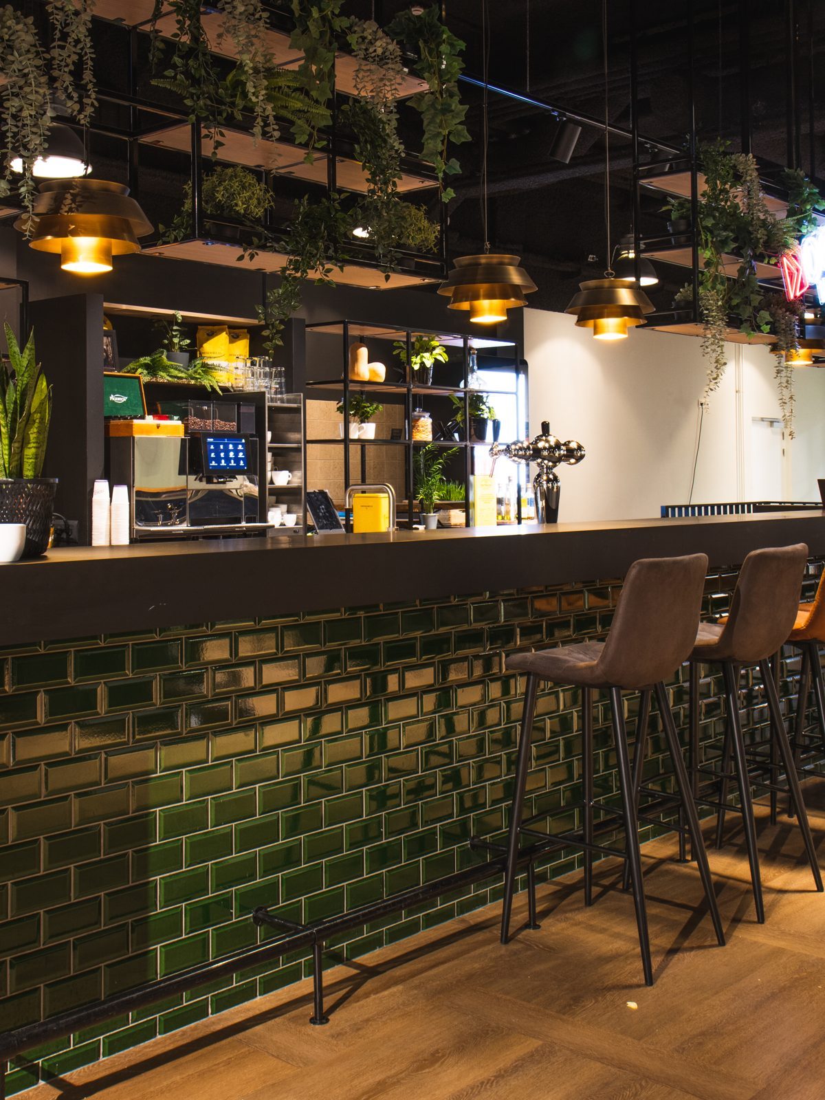 De bar bij OGD Delft met donkergroene tegels, houten barkrukken, warme verlichting en hangende planten.