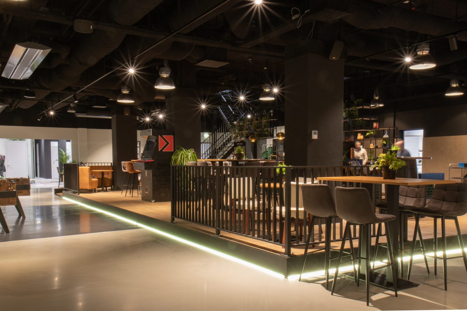 De gedeelde ruimte met verhoogde houten vloer, zwarte balustrades, tafels, stoelen en open donkere plafonds.