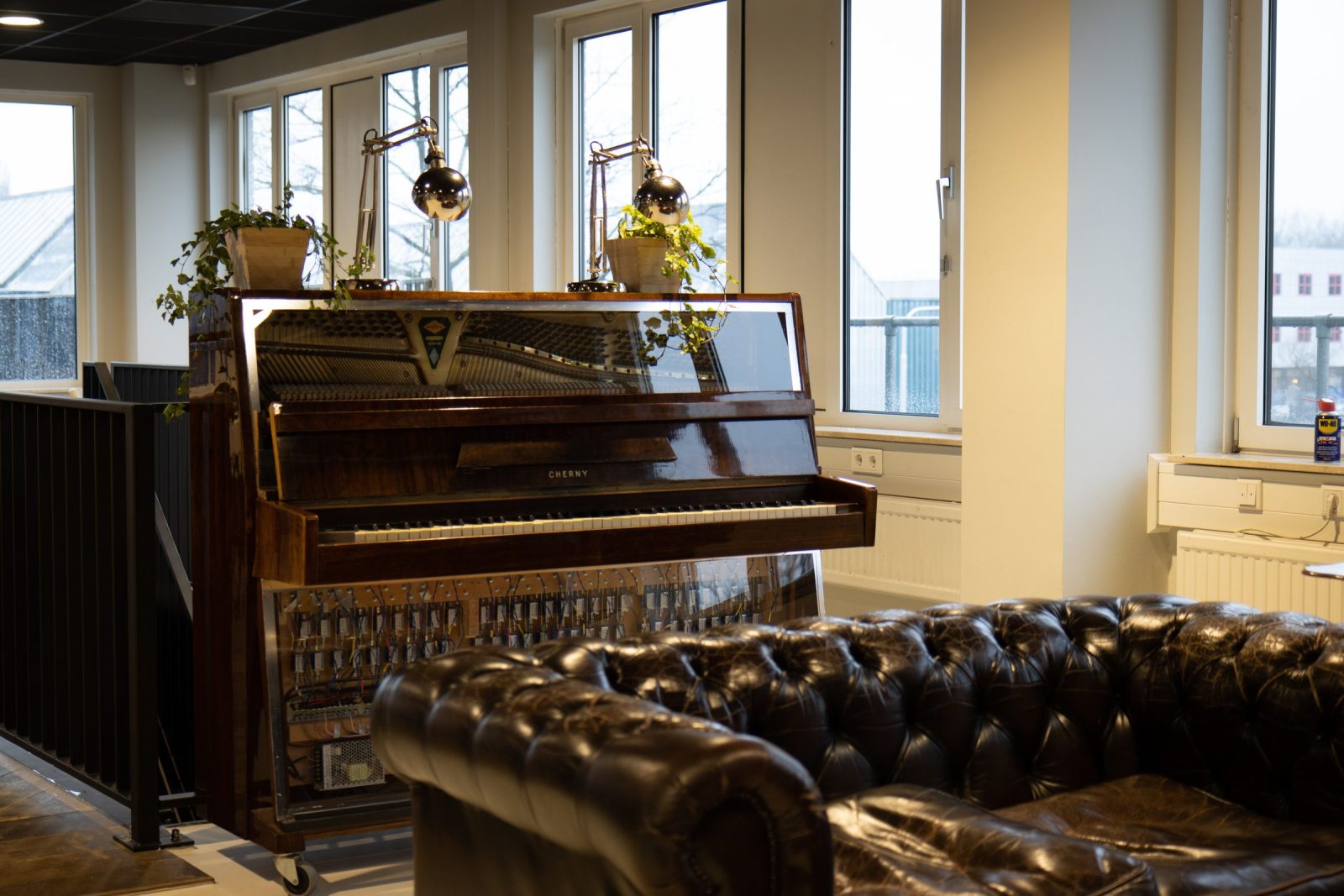 Een houten piano achter een donkerleren Chesterfield-bank, bij hoge ramen en planten.