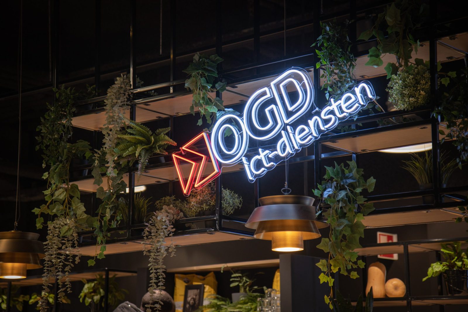Het lichtgevende OGD ict-diensten-logo boven de bar, tussen houten plantenbakken en hanglampen.