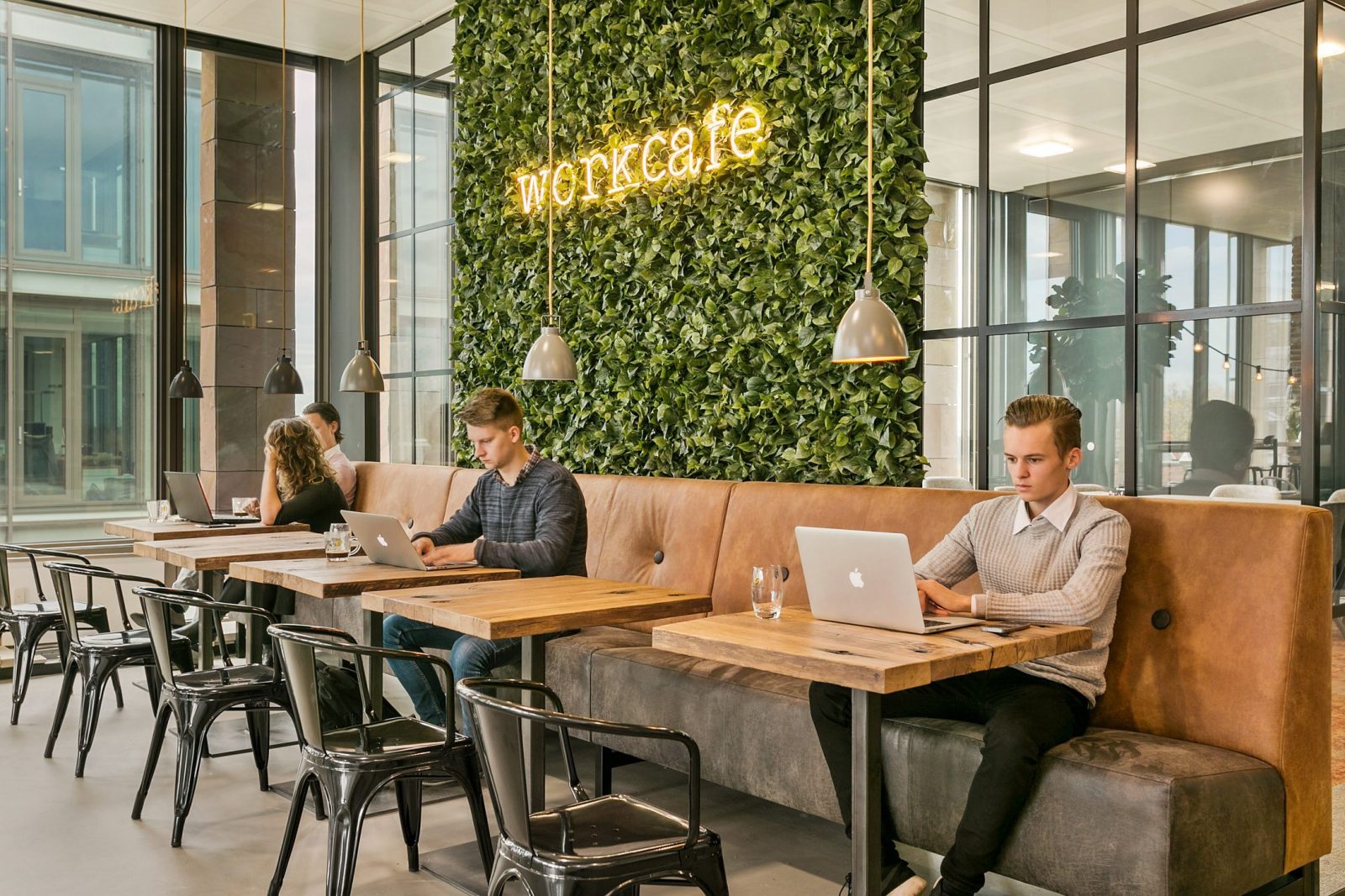 Het workcafé met een groene plantenwand, een cognackleurige bank, lange tafels en mensen met laptops.
