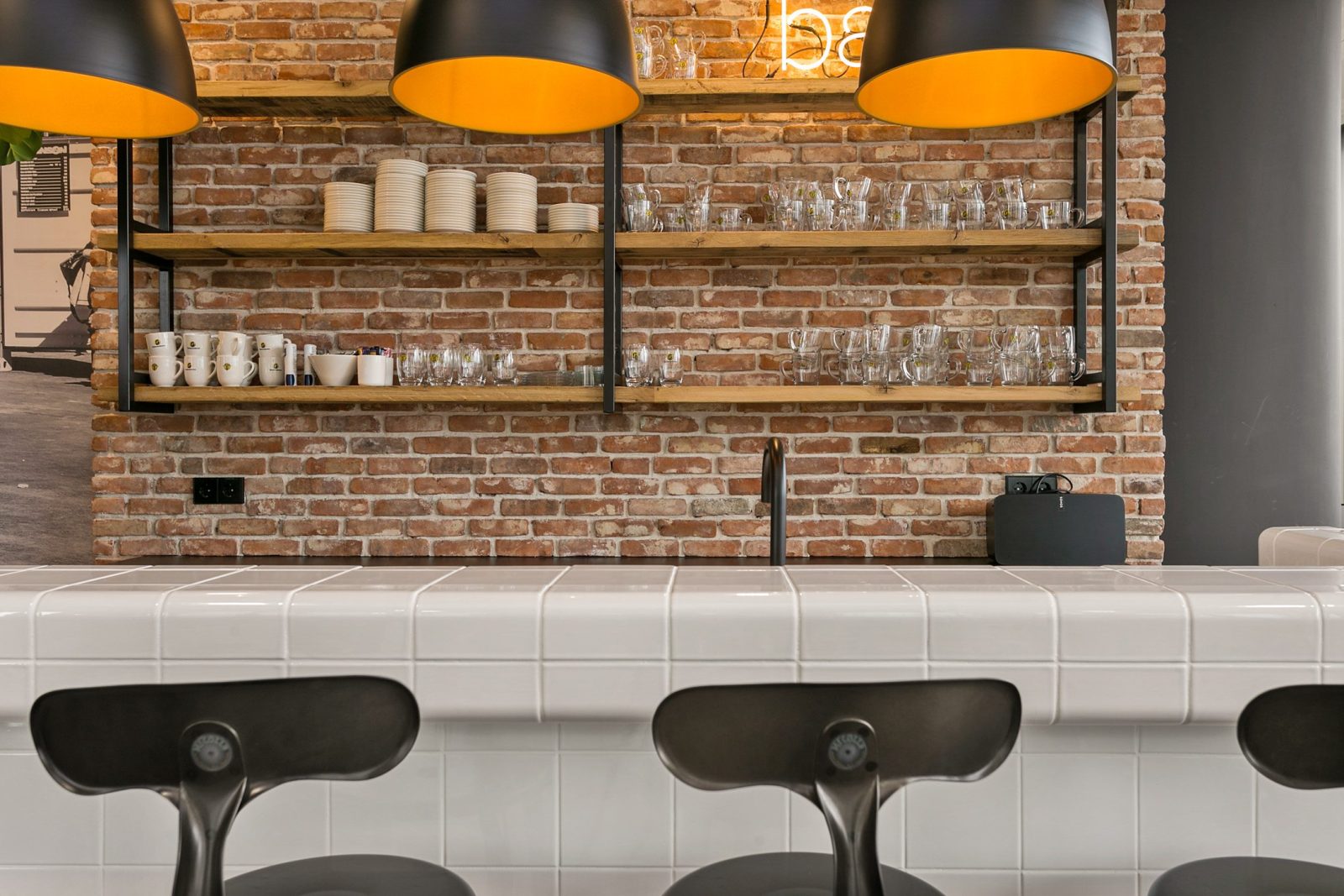 Een witte betegelde bar voor een bakstenen wand, met zwarte barkrukken en donkere lampen met een gele binnenkant.