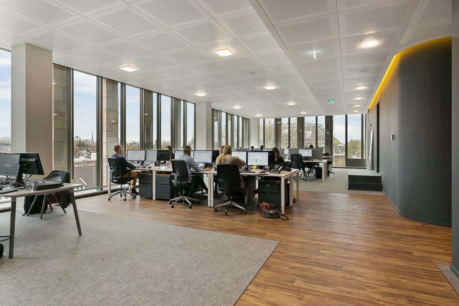 Een open werkvloer met bureaus, hoge ramen, een houten looproute en een donkere wand met een geel lichtaccent.
