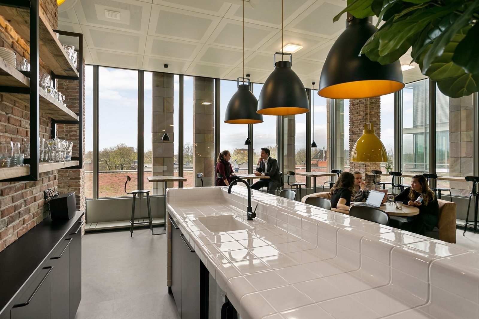 Een witte bar onder zwarte hanglampen, met hoge ramen en het workcafé op de achtergrond.