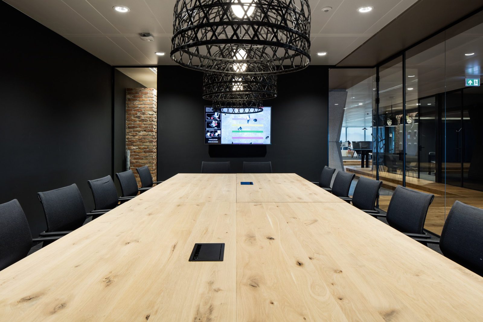 Een lange houten vergadertafel met donkere stoelen onder grote zwarte gevlochten hanglampen, naast glazen wanden.