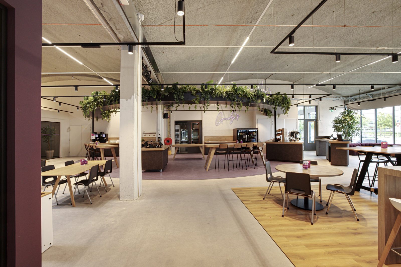 De gezamenlijke caféruimte met houten tafels, een koffiebar, planten en een bordeauxrode wand.