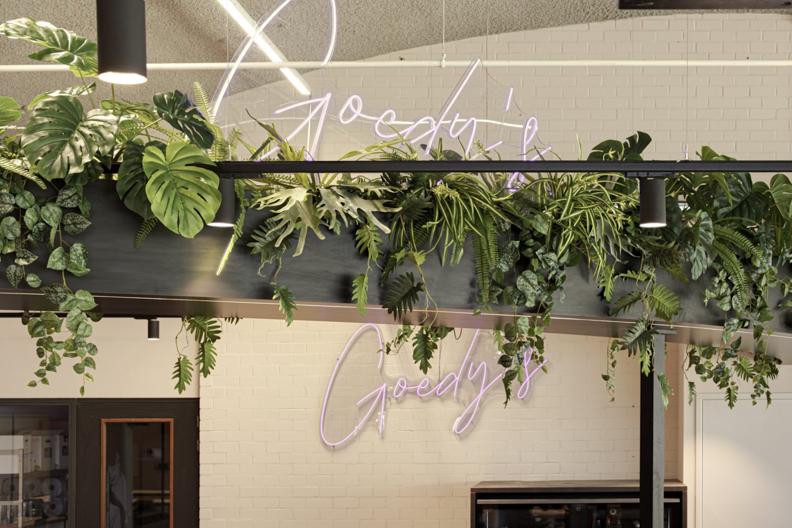 Hangende planten en zwarte spots boven de koffiebar, met een lichtgevend Goody’s-logo op de witte bakstenen wand.