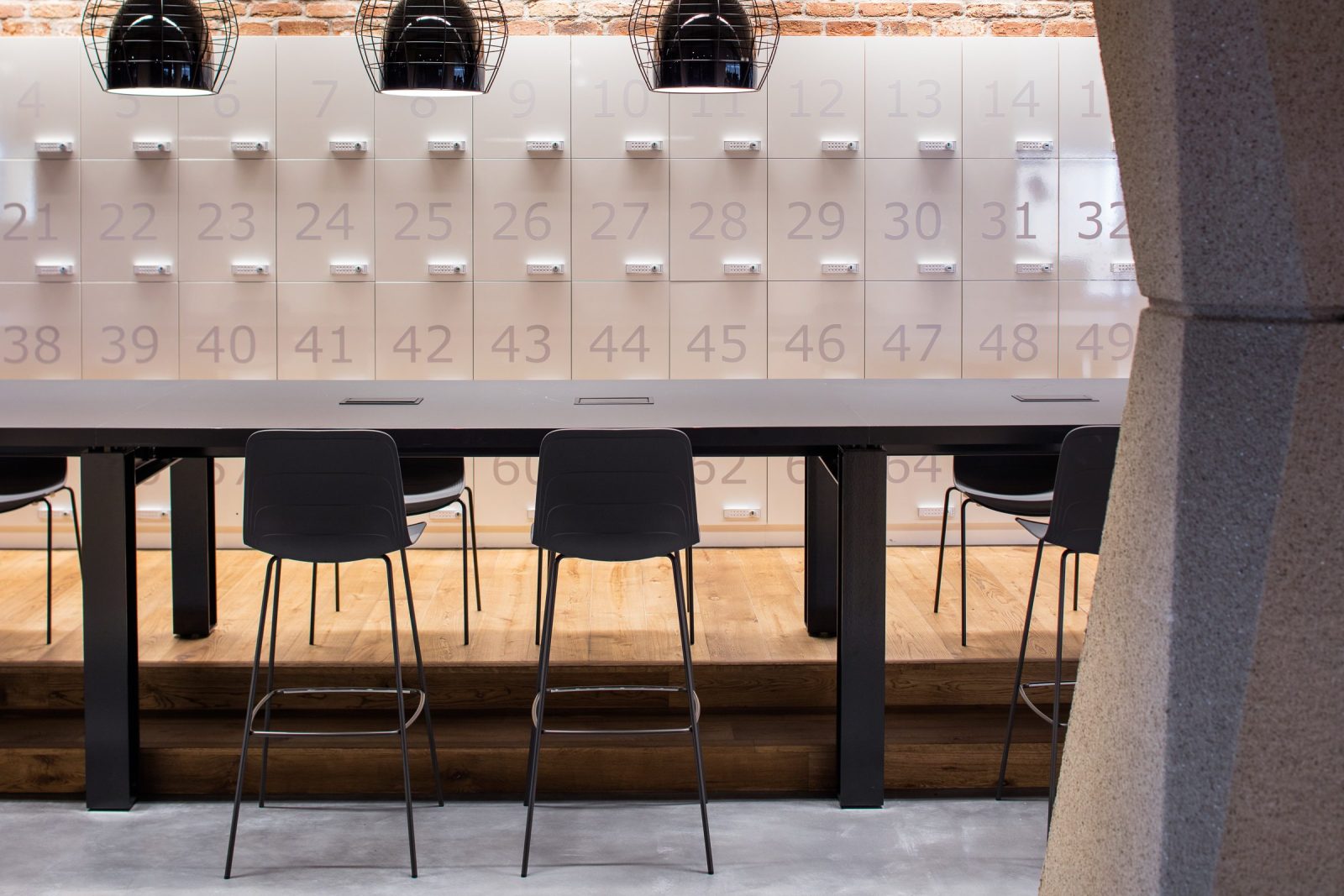 Een hoge tafel met zwarte stoelen voor een witte wand met genummerde vakken en een bakstenen rand.