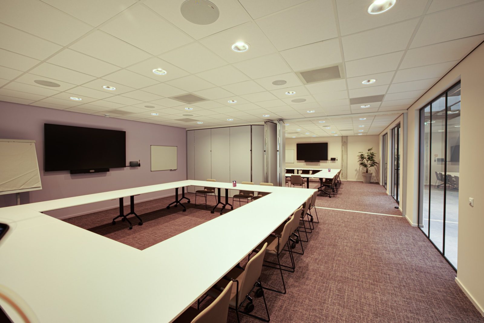 Een grote vergaderruimte met tafels in een U-opstelling, bordeauxrode stoelen en een wandbeeldscherm.