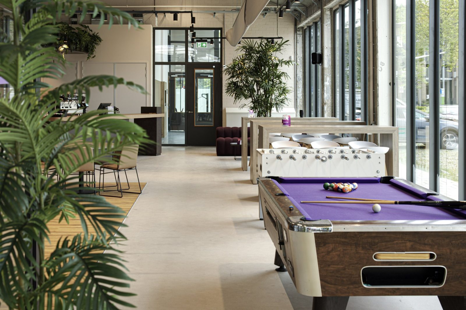 Een pooltafel en tafelvoetbal naast hoge ramen, met planten en zitplekken op de achtergrond.