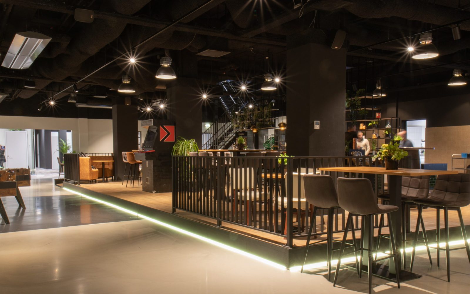 Een brede blik op de ontmoetingsruimte bij OGD Delft, met zwarte kolommen, een donkere bar, houten tafels en planten.