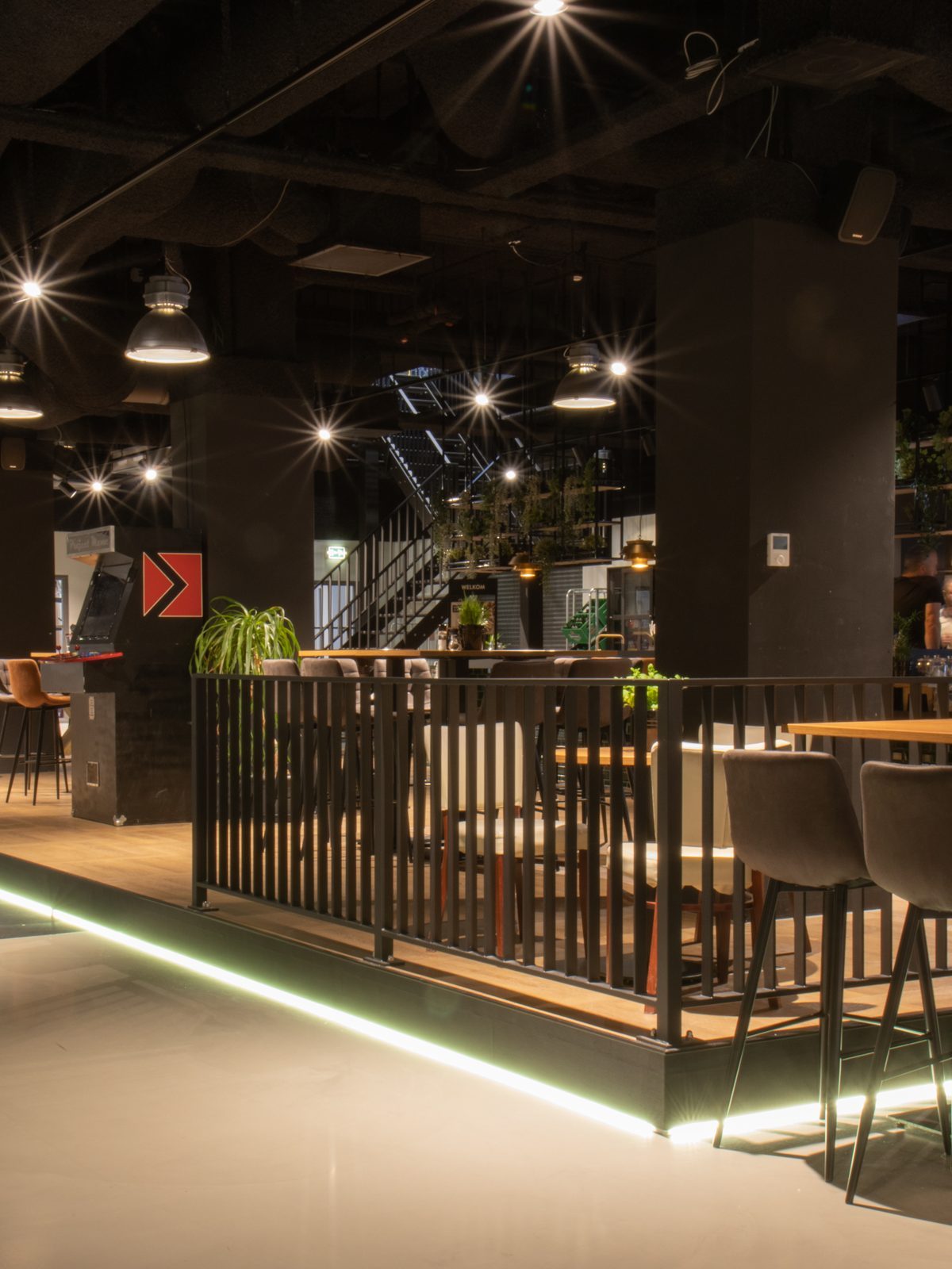 Een brede blik op de ontmoetingsruimte bij OGD Delft, met zwarte kolommen, een donkere bar, houten tafels en planten.